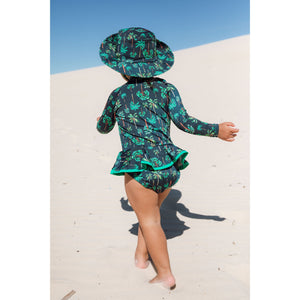 Bebe de 2 anos está vestindo maiozinho de praia infantil feminino com manga longa, babadinho, e zipper na frente, estampa coqueiros azul marinho com chapéuzinho combinando, da Lili Sampedro