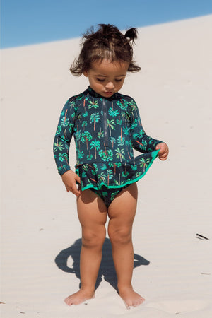 Bebe está vestindo maiozinho de praia infantil de manga longa com babadinhos e zipper na frente, na estampa coqueiros azul marinho, com protecao UV da Lili Sampedro