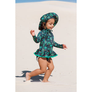 Bebe de 2 anos está vestindo maiozinho de praia infantil feminino com manga longa, babadinho, e zipper na frente, estampa coqueiros azul marinho com chapéuzinho combinando, da Lili Sampedro