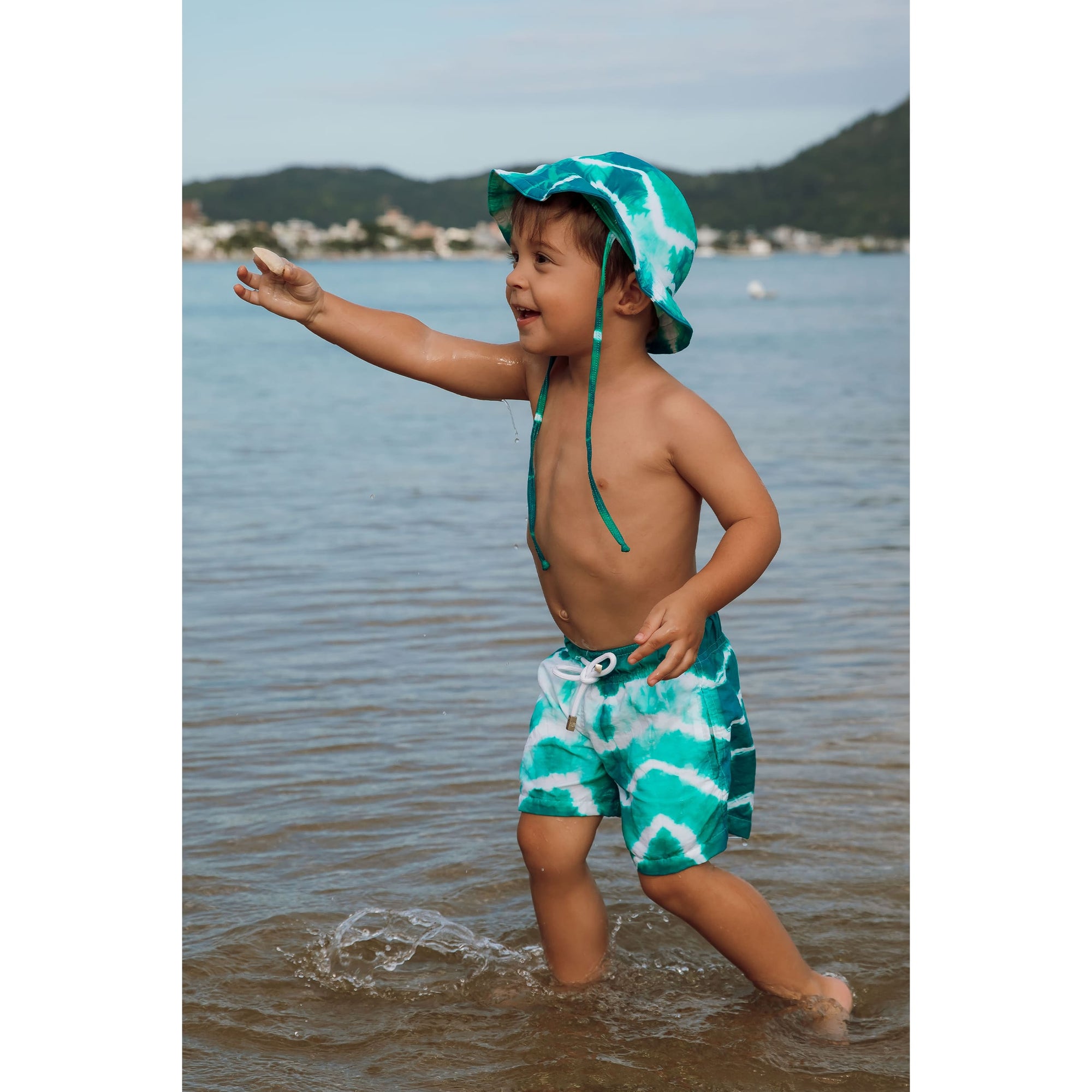 Menino de 2 anos vestindo short infantil de praia Tie Dye Verde com chapéuzinho infantil combinando da Lili Sampedro