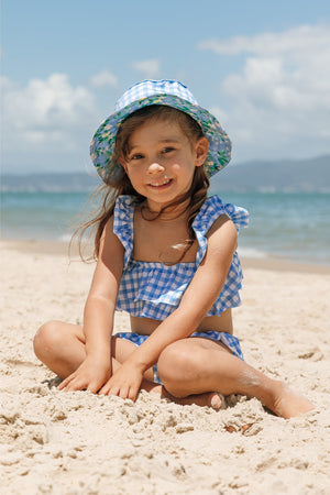 Menina na praia sentada com biquíni infantil na estampa Vichy e chapéuzinho de praia com protecao UV na mesma estampa combinando da Lili Sampedro