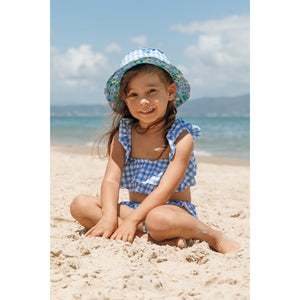 Menina na praia sentada com biquíni infantil na estampa Vichy e chapéuzinho de praia com protecao UV na mesma estampa combinando da Lili Sampedro